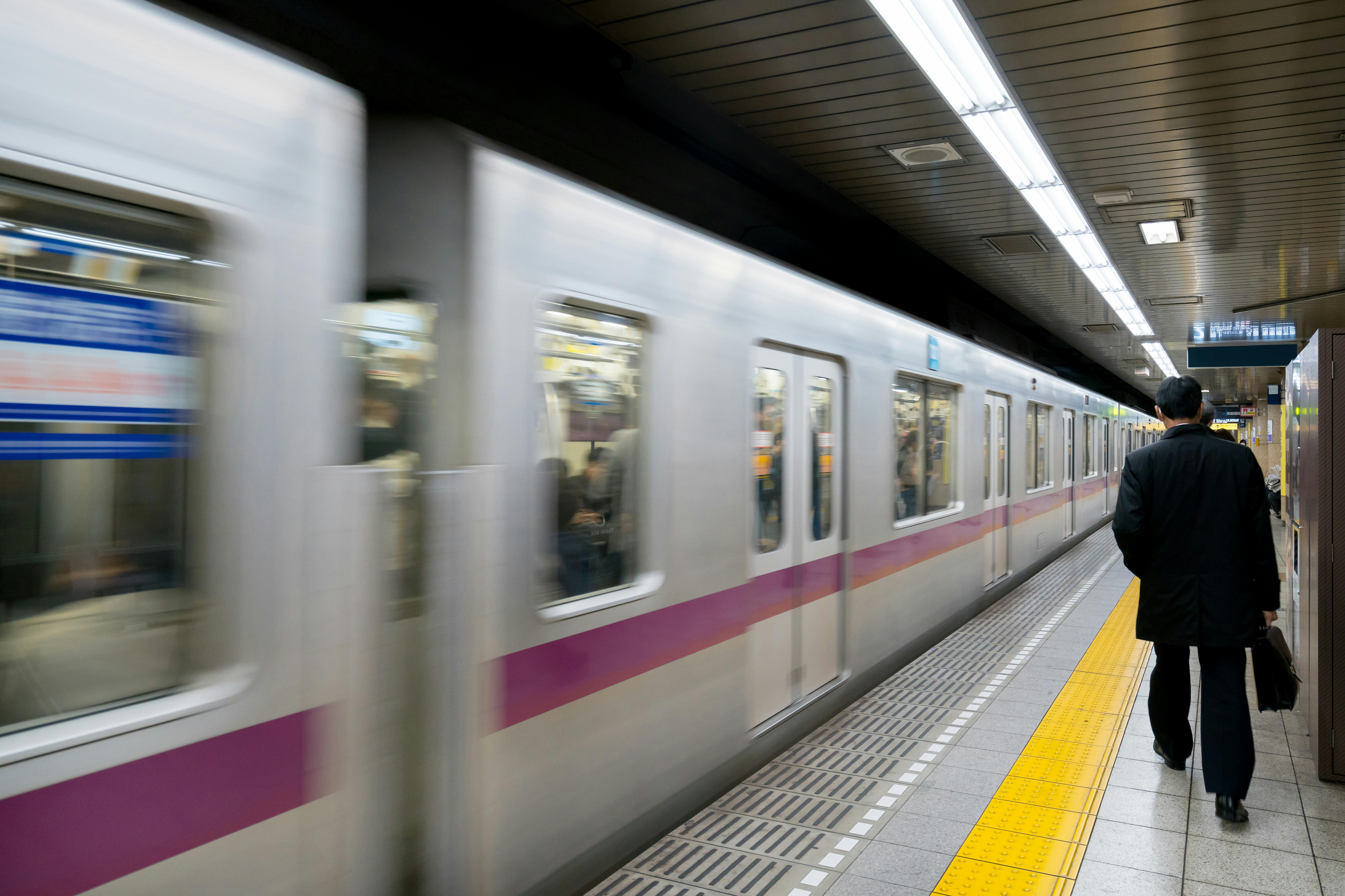 A man in a suit walks along a subway platform as a train speeds by, with blurred motion. The platform has yellow tactile paving for the visually impaired, and fluorescent lights line the ceiling.