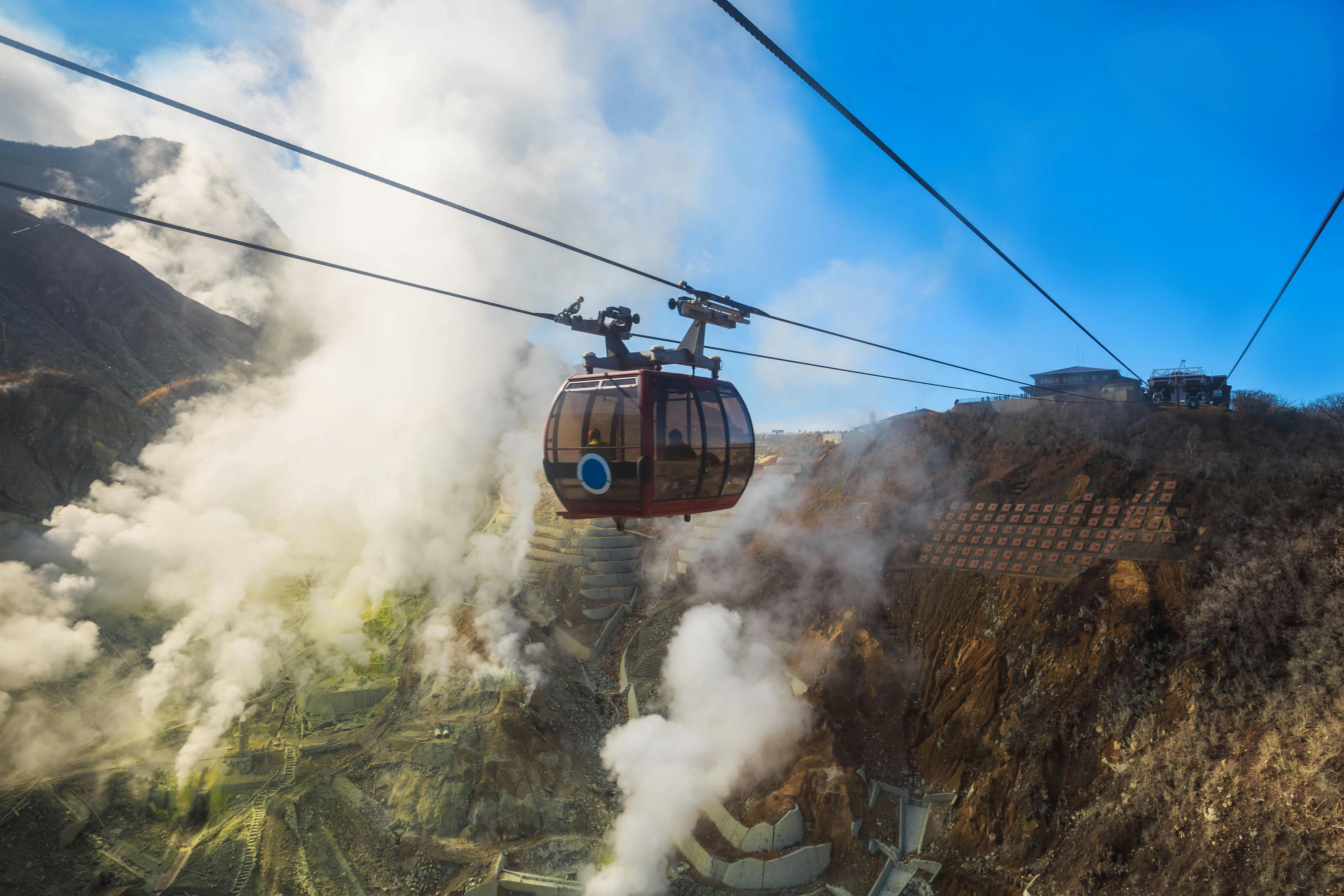 A cable car travels over a steaming, rocky mountainside on a bright day, with clouds of white steam rising from the ground and a clear blue sky overhead.