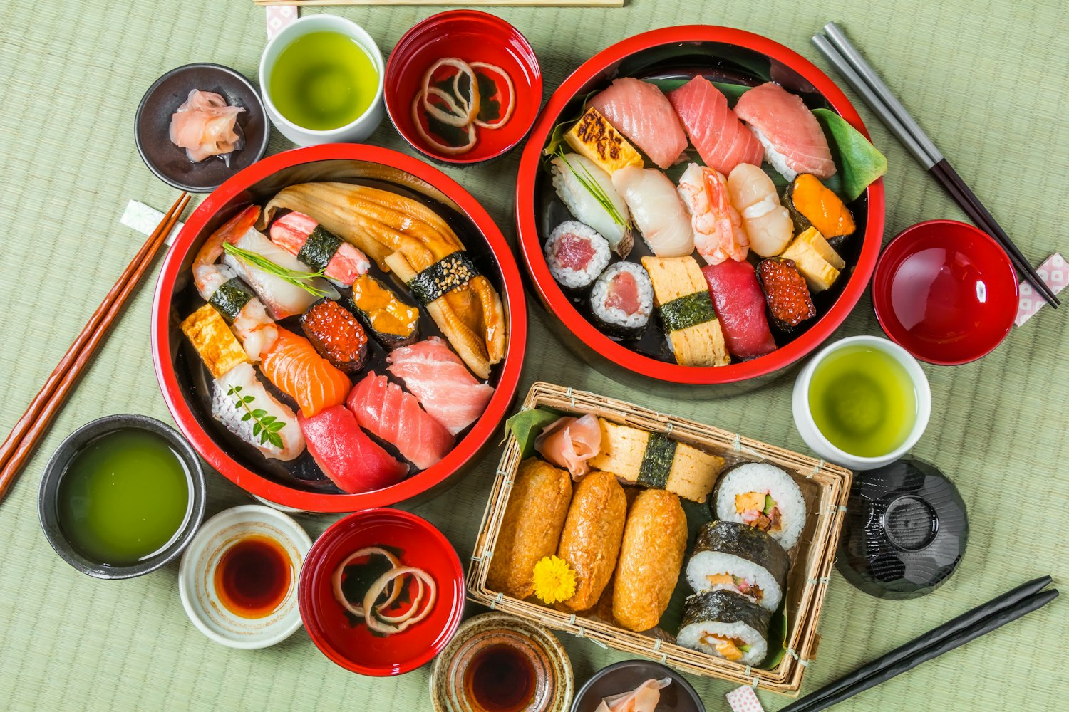 Two red plates filled with assorted sushi, including nigiri and sashimi, are on a table. A small wooden tray holds sushi rolls and inari. Soy sauce, ginger, wasabi, and green tea cups are also visible, creating a colorful Japanese meal setting.