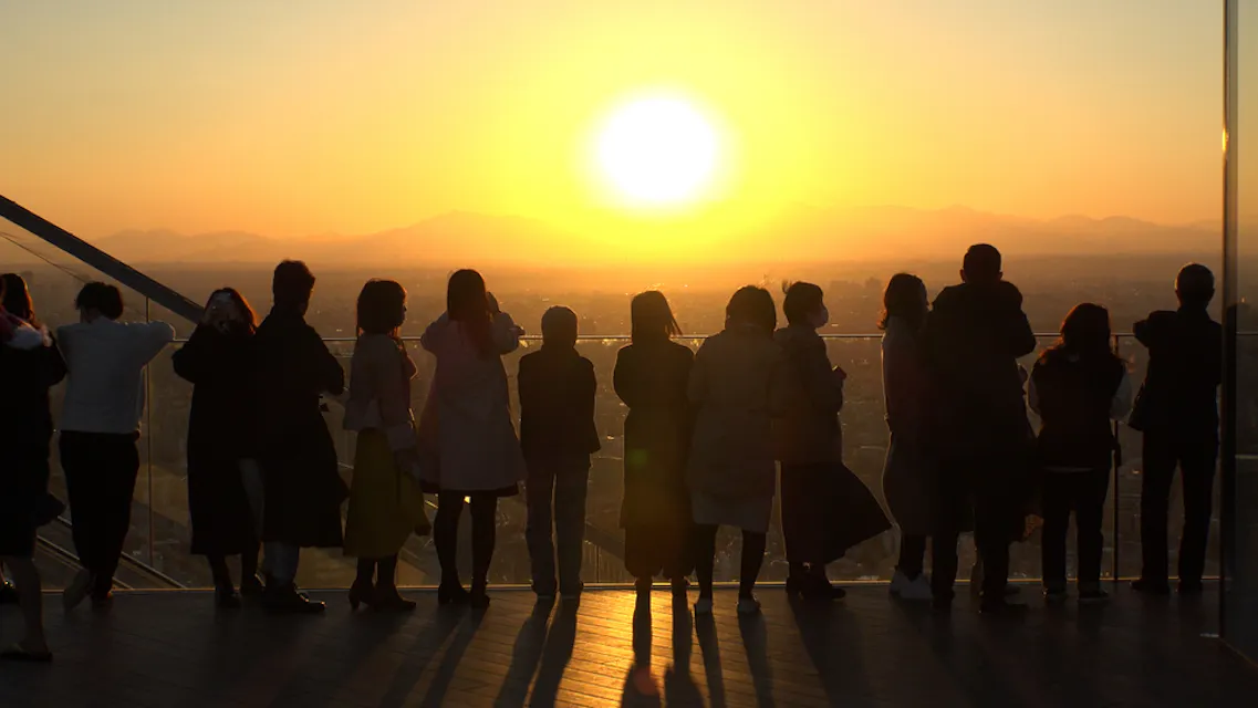 Sunset from the Shibuya Sky rooftop, the most popular time slot of the day Visitors stand in silhouette along the glass edge of the Shibuya Sky rooftop as the sun sets over western Tokyo
