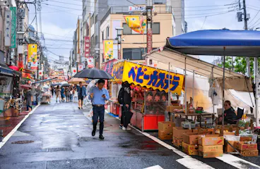 People walk through a lively outdoor market on a rainy day in Japan, some holding umbrellas, with colorful stalls selling various goods and produce lining both sides of the wet street.