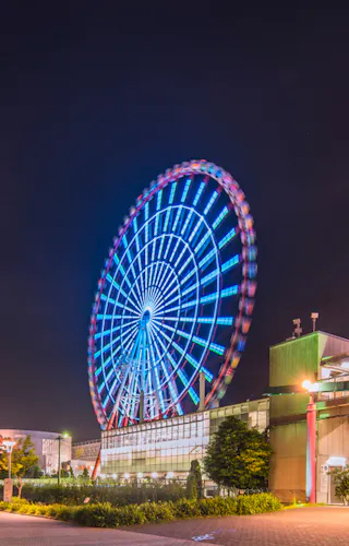 Odaiba Illuminated Palette Town Ferris Wheel A vibrant Ferris wheel lit up at night, located near a building with a sign reading "Palette Town" in colorful letters. The scene includes a well-lit pathway and surrounding greenery, set against a dark sky.