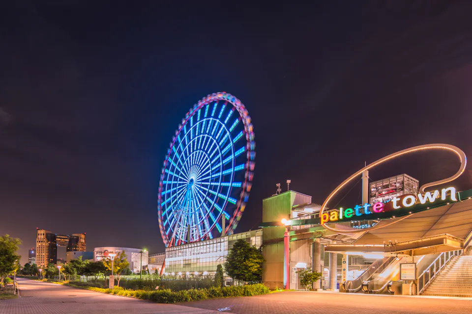 A vibrant Ferris wheel lit up at night, located near a building with a sign reading "Palette Town" in colorful letters. The scene includes a well-lit pathway and surrounding greenery, set against a dark sky.