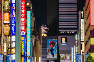 Colorful neon signs in Japanese line a busy street at night in Tokyo. A large Godzilla head is visible atop a building, next to a Hotel Gracery and a billboard featuring Mario from Super Mario Bros.