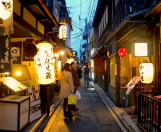 People walk down a narrow, lantern-lit street lined with traditional Japanese buildings and restaurants in the evening. Bright signs and paper lanterns illuminate the scene, creating a warm, inviting atmosphere.