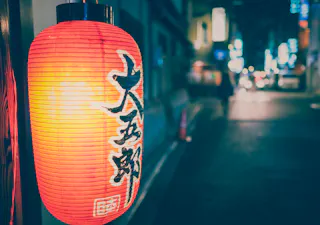 A red Japanese paper lantern with black kanji characters hangs outside, illuminating a city street at night with blurred lights in the background.