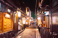 A narrow, lantern-lit alleyway lined with traditional wooden buildings at night, likely in Japan. The street is empty except for a few blurred figures in the distance, creating a warm and inviting atmosphere.