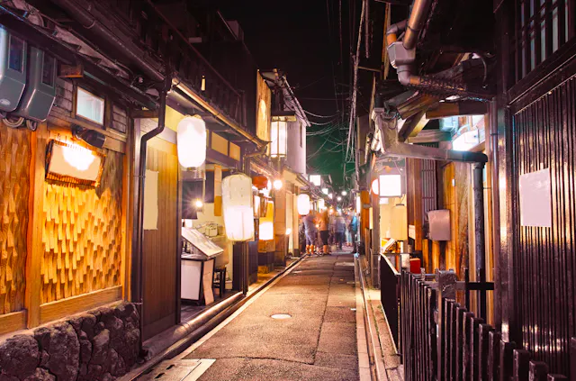 A narrow, lantern-lit alleyway lined with traditional wooden buildings at night, likely in Japan. The street is empty except for a few blurred figures in the distance, creating a warm and inviting atmosphere.