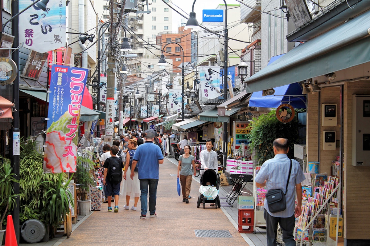 Yanaka Ginza Shopping Street A bustling street scene in a Japanese shopping district. People walk along the narrow street lined with shops displaying colorful signs and awnings. A man pushes a stroller, and various goods are visible in the shop windows.