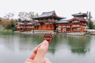 A hand holds a Japanese coin in front of the Byodo-in Temple, with the temple’s ornate red buildings and a reflective pond in the background.