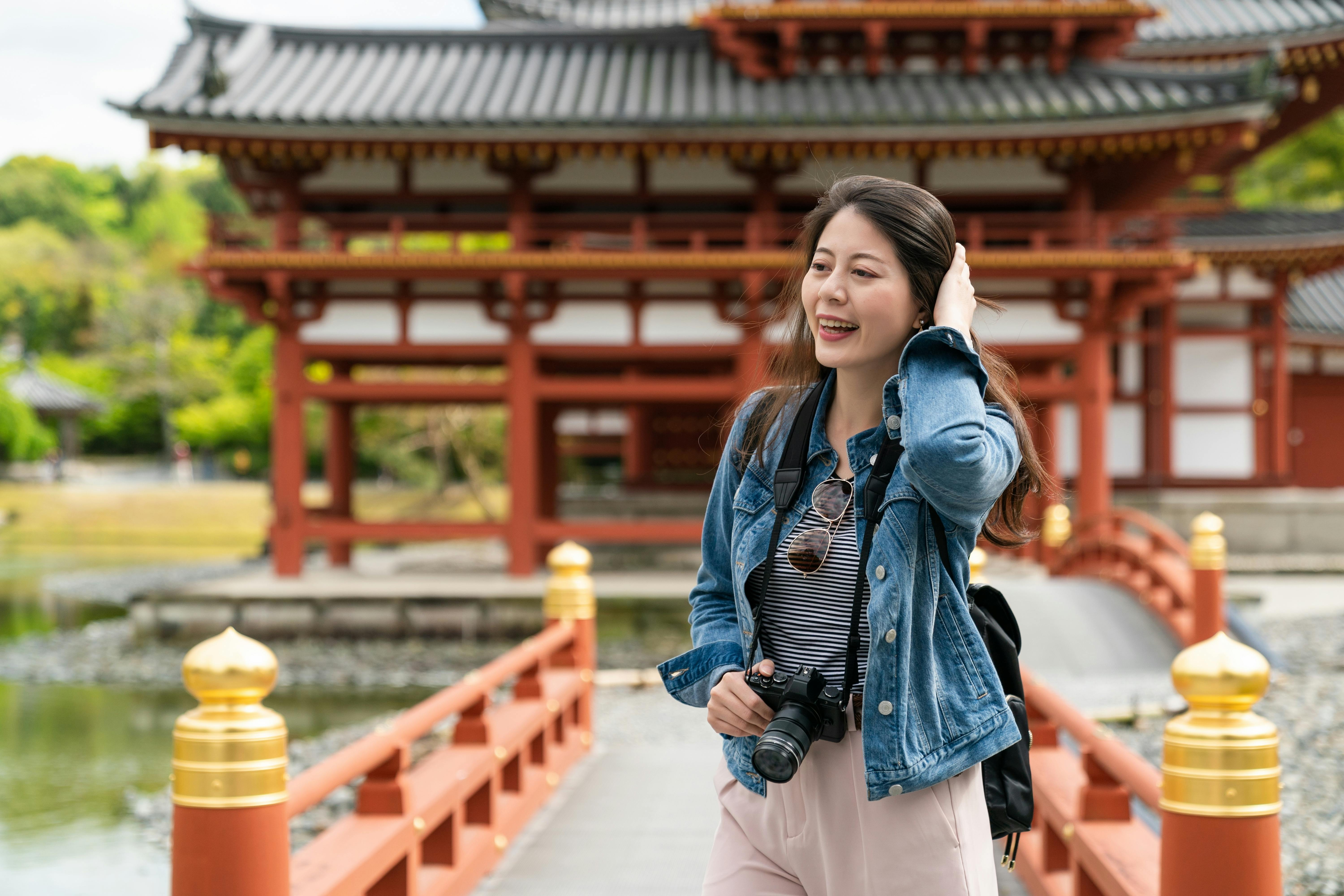 A young woman with a camera around her neck stands on a red bridge, smiling and touching her hair, with a traditional Japanese temple and pond in the background.