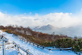 A snowy path bordered by a wooden fence overlooks a calm lake with tree-covered islands and distant mountains under a partly cloudy blue sky.