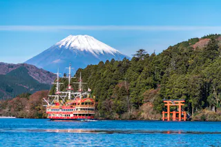 A scenic view of a pirate-themed boat sailing on a lake, with a red torii gate and lush green forest in the background. Snow-capped Mount Fuji rises majestically under a clear blue sky.