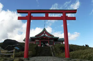 A large red torii gate stands at the entrance of a traditional Japanese shrine with green roofs, set atop stone steps under a partly cloudy sky. Grass and rope-lined paths flank the steps.