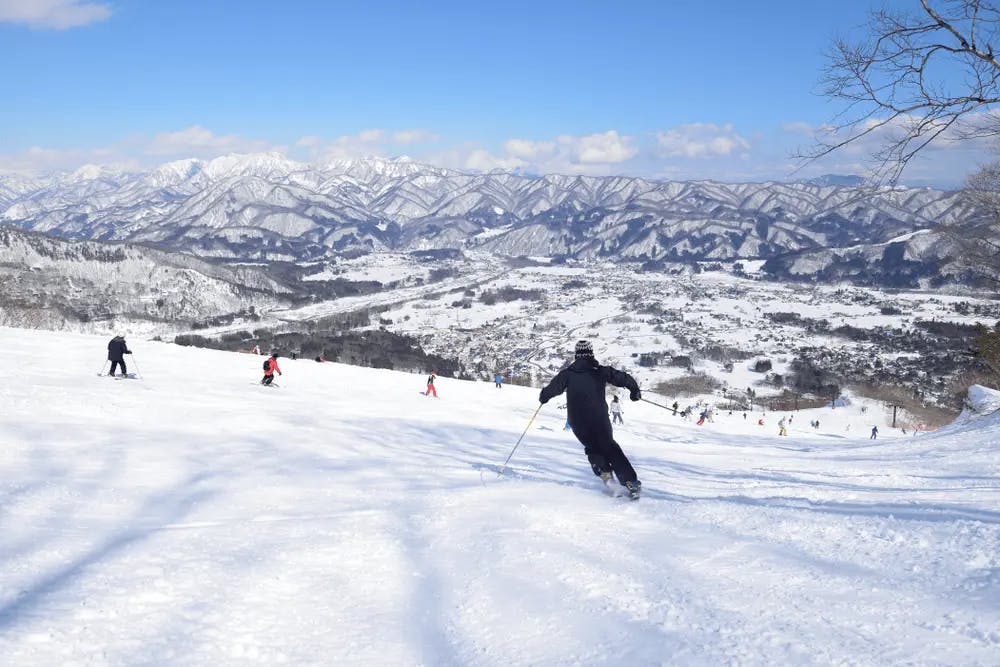 A skier in a black outfit descends a snow-covered slope under a clear blue sky. Several other skiers can be seen in the background, along with a vast, mountainous landscape covered in snow. The valley below is dotted with structures and trees, adding to the scenic beauty.