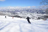 A skier in a black outfit descends a snow-covered slope under a clear blue sky. Several other skiers can be seen in the background, along with a vast, mountainous landscape covered in snow. The valley below is dotted with structures and trees, adding to the scenic beauty.