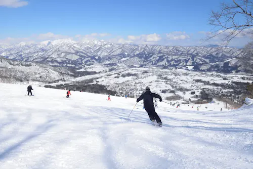 A skier in a black outfit descends a snow-covered slope under a clear blue sky. Several other skiers can be seen in the background, along with a vast, mountainous landscape covered in snow. The valley below is dotted with structures and trees, adding to the scenic beauty.