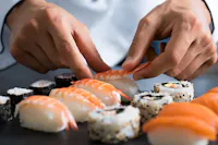 Close-up of a chef's hands expertly preparing assorted sushi. The selection includes nigiri topped with shrimp and various types of rolled sushi with ingredients like fish, seaweed, and rice, all displayed on a dark slate surface.