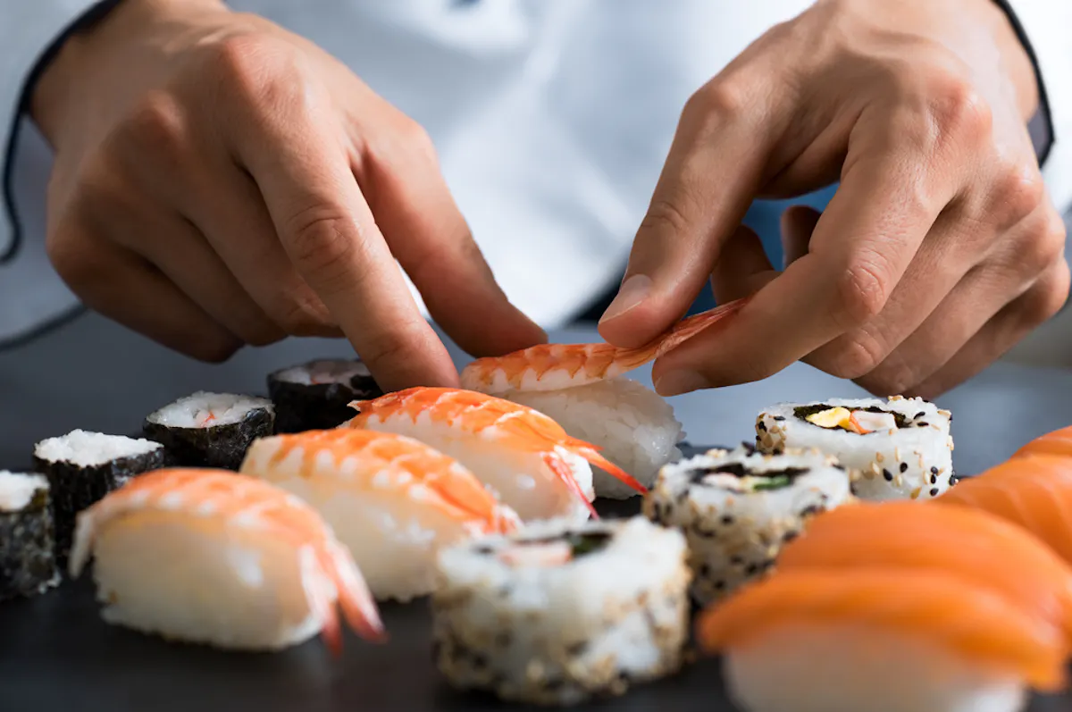 Close-up of a chef's hands expertly preparing assorted sushi. The selection includes nigiri topped with shrimp and various types of rolled sushi with ingredients like fish, seaweed, and rice, all displayed on a dark slate surface.