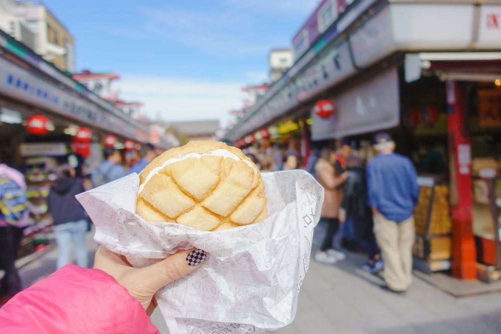 A hand holding a melon pan bread wrapped in paper, with a busy outdoor market and people walking in the background on a sunny day.