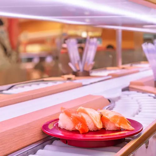 Conveyor Belt Sushi Restaurants A plate with sushi pieces, including salmon and shrimp nigiri, is on a conveyor belt in a sushi restaurant. The background has a blurred interior with chopsticks in containers and a warm, inviting setting.