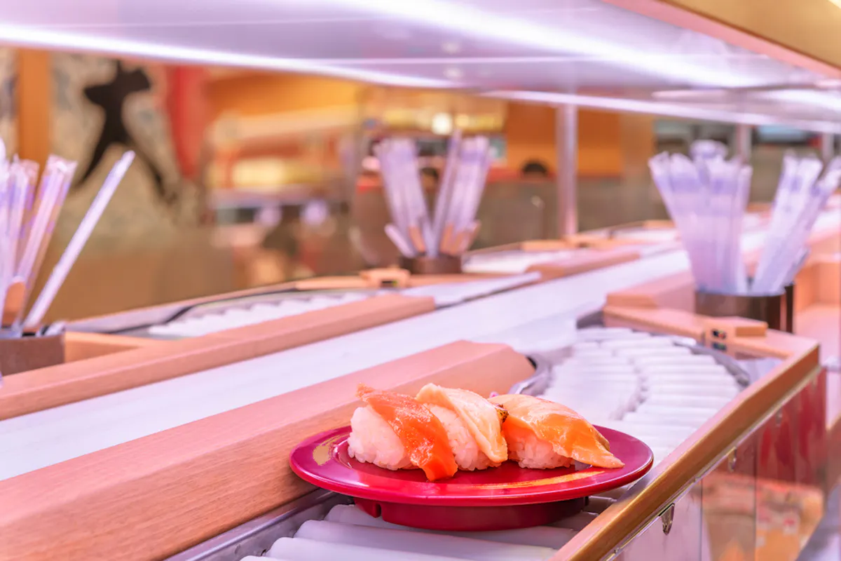 Conveyor Belt Sushi Restaurants A plate with sushi pieces, including salmon and shrimp nigiri, is on a conveyor belt in a sushi restaurant. The background has a blurred interior with chopsticks in containers and a warm, inviting setting.