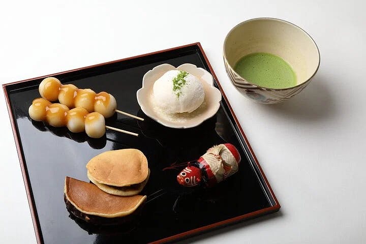 A tray with Japanese sweets: two skewers of dango, a round white mochi with green tea powder, two small dorayaki pancakes, and a daruma doll, beside a bowl of green matcha tea on a white background.