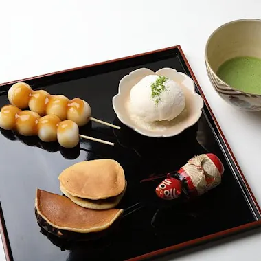 Wagashi Cooking Class A tray with Japanese sweets: two skewers of dango, a round white mochi with green tea powder, two small dorayaki pancakes, and a daruma doll, beside a bowl of green matcha tea on a white background.