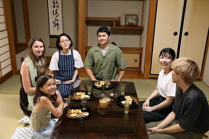 Six people sit around a low table with snacks and drinks in a traditional Japanese-style room, smiling and posing for the photo. The room features tatami mats, sliding doors, and decorative calligraphy.