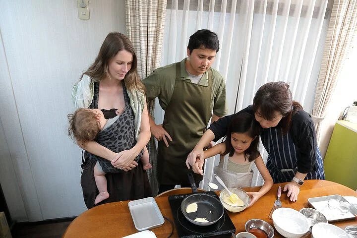 A woman holding a baby stands beside a man in an apron as an older woman helps a young girl pour batter into a pan on a stovetop, all gathered around a kitchen table preparing food together.