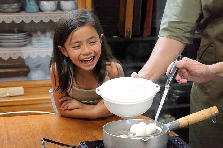 A young girl smiles excitedly at a pot on a stove while an adult uses a ladle to scoop round dumplings into a white colander. The scene takes place in a cozy kitchen with shelves of dishes in the background.