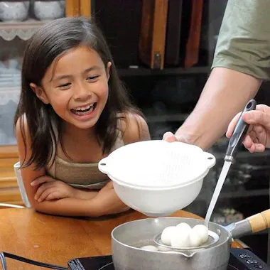 Wagashi Cooking Class A young girl smiles excitedly at a pot on a stove while an adult uses a ladle to scoop round dumplings into a white colander. The scene takes place in a cozy kitchen with shelves of dishes in the background.