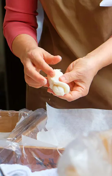 Wagashi Cooking Class A person wearing a brown apron and a pink long-sleeve shirt shapes a piece of dough with their hands over a work surface covered with parchment paper.