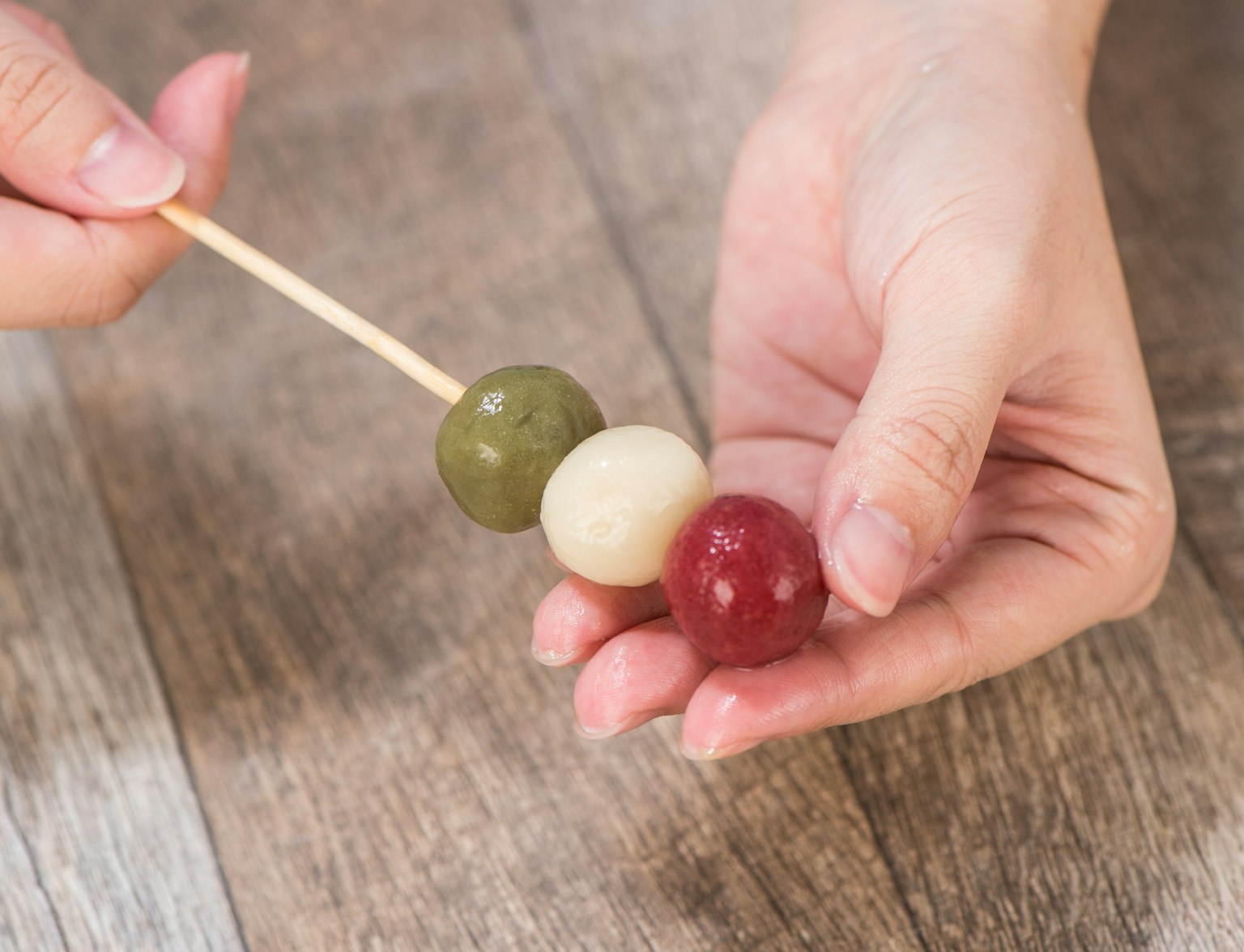Wagashi Cooking Class A person holding a skewer with three colored dango balls—green, white, and red—over a wooden surface.