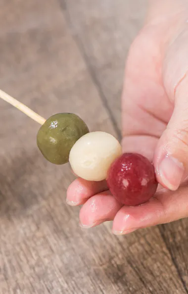 Wagashi Cooking Class A person holding a skewer with three colored dango balls—green, white, and red—over a wooden surface.