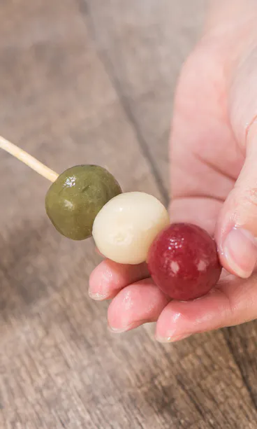 Wagashi Cooking Class A person holding a skewer with three colored dango balls—green, white, and red—over a wooden surface.