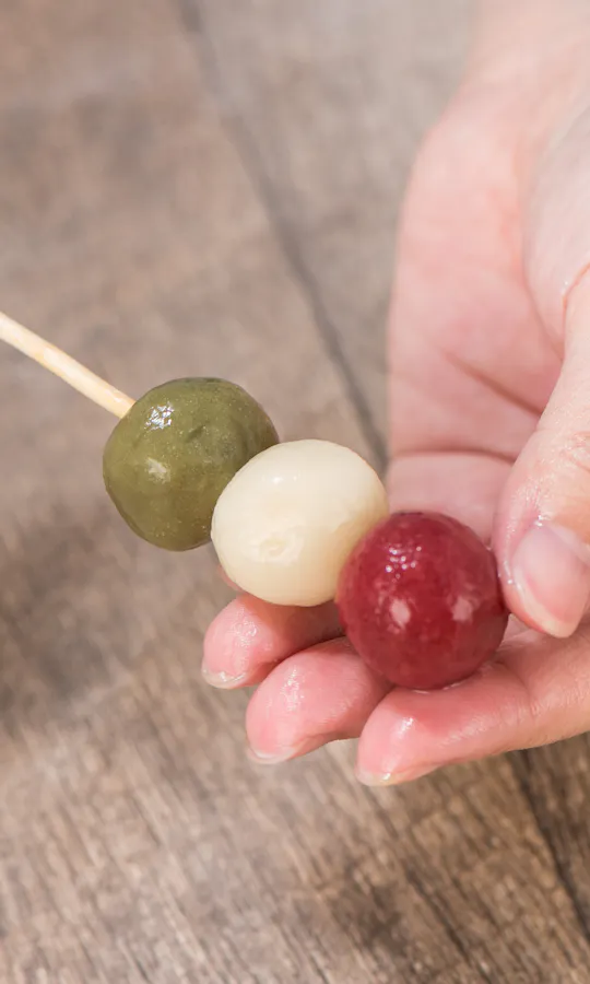 Wagashi Cooking Class A person holding a skewer with three colored dango balls—green, white, and red—over a wooden surface.