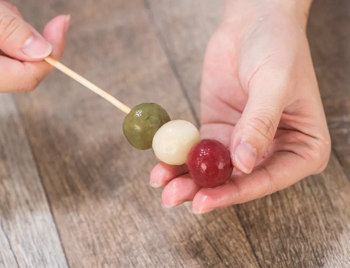 Wagashi Cooking Class A person holding a skewer with three colored dango balls—green, white, and red—over a wooden surface.