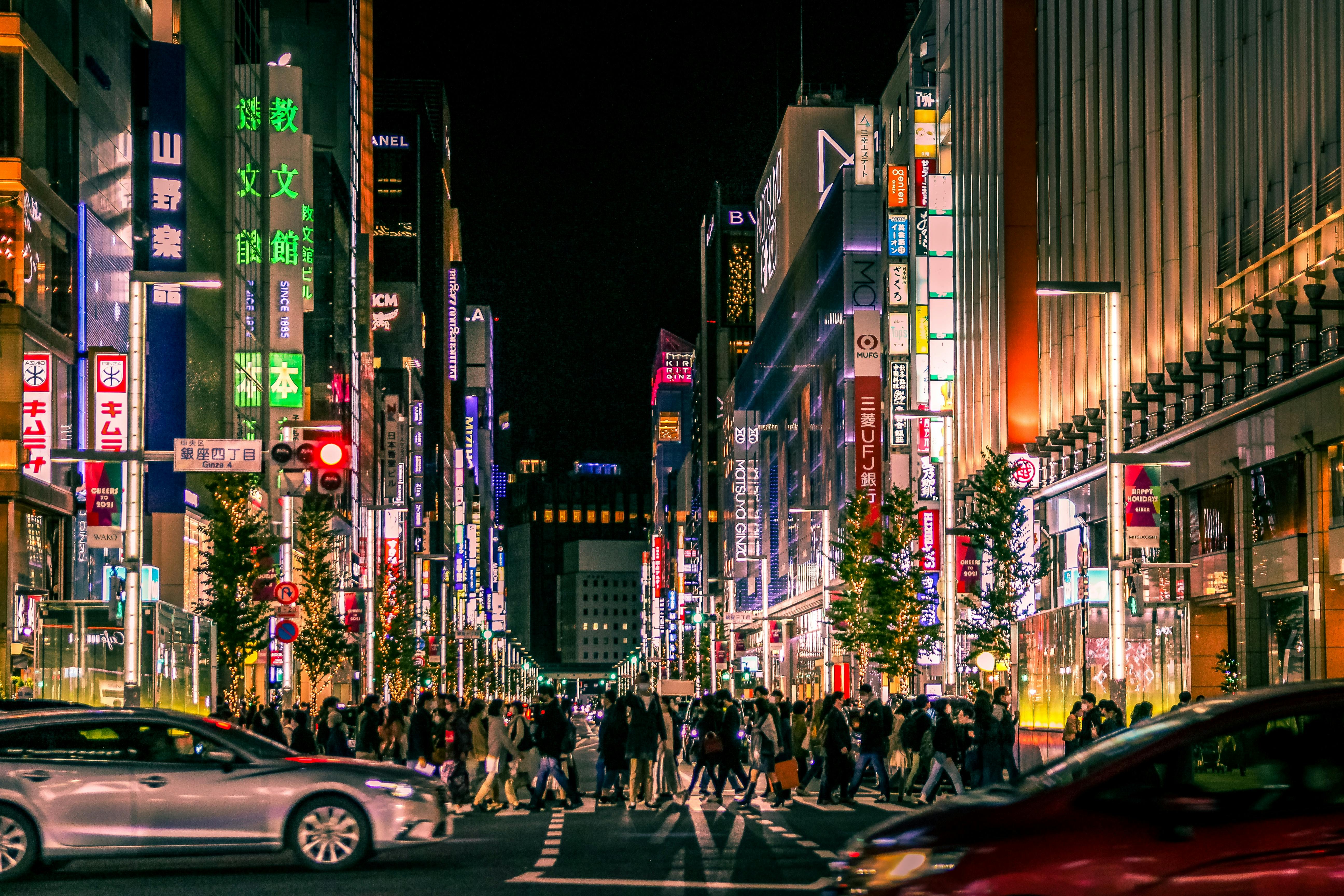 A busy city street at night, filled with people crossing and cars passing by, surrounded by tall buildings brightly lit with colorful neon signs and advertisements in Japanese.