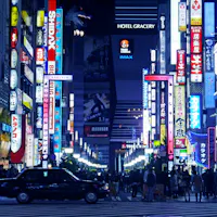 Shinjuku A busy city street at night in Tokyo’s Shinjuku district, filled with colorful neon signs and billboards in Japanese. Crowds of people walk along the sidewalks and a black taxi waits at a crosswalk.