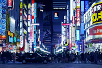 A busy city street at night in Tokyo’s Shinjuku district, filled with colorful neon signs and billboards in Japanese. Crowds of people walk along the sidewalks and a black taxi waits at a crosswalk.