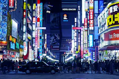 A busy city street at night in Tokyo’s Shinjuku district, filled with colorful neon signs and billboards in Japanese. Crowds of people walk along the sidewalks and a black taxi waits at a crosswalk.