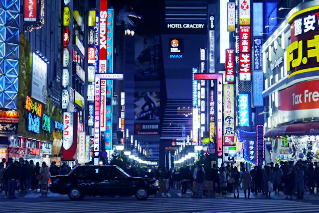 A busy city street at night in Tokyo’s Shinjuku district, filled with colorful neon signs and billboards in Japanese. Crowds of people walk along the sidewalks and a black taxi waits at a crosswalk.