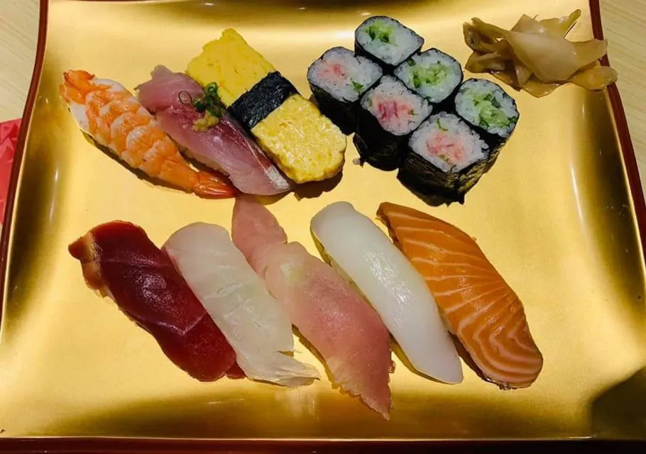 一盘金色的盘子上摆满了各种寿司，包括各种配料的握寿司，如金枪鱼、三文鱼、虾和鸡蛋。旁边还有六块黄瓜卷和一份腌姜。