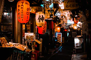 A narrow, dimly lit alleyway in Japan at night, lined with glowing red and yellow lanterns, Japanese signs, and small restaurants with menus displayed outside. The scene feels lively and inviting.