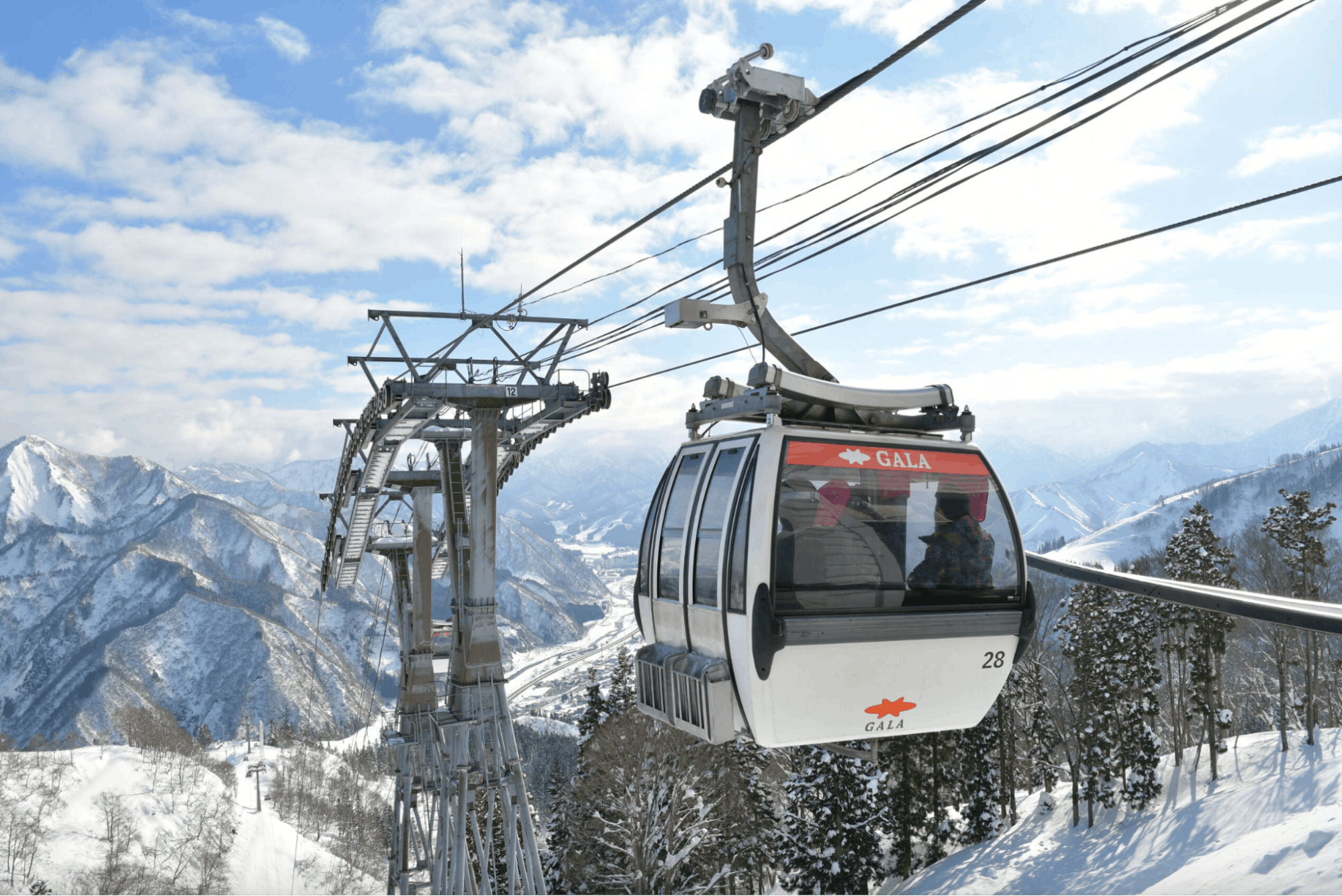 A cable car travels up a snowy mountain with snow-covered peaks and trees in the background under a partly cloudy sky. The cable car is labeled "GALA.