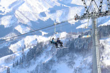Two people sit on a ski lift ascending a snowy mountain, surrounded by snow-covered trees and slopes under a clear sky. A single empty chair hangs nearby.