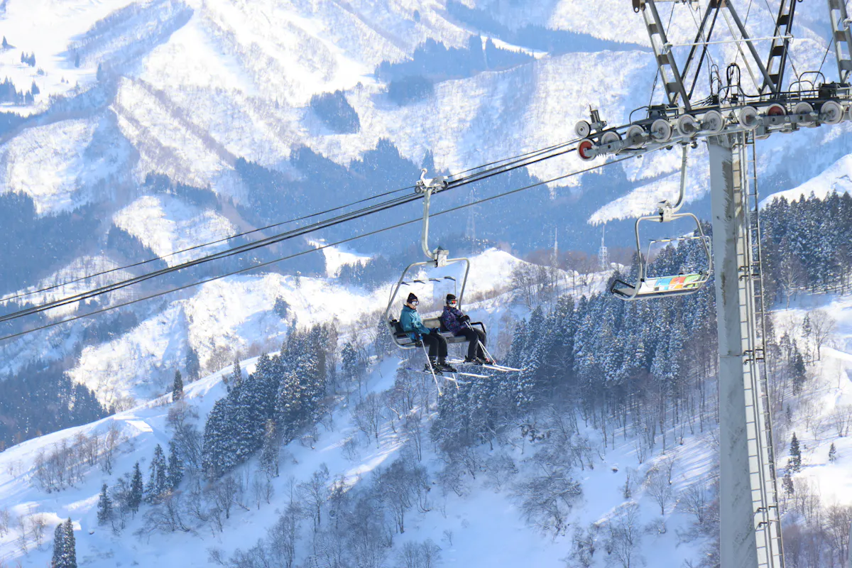 Two people sit on a ski lift ascending a snowy mountain, surrounded by snow-covered trees and slopes under a clear sky. A single empty chair hangs nearby.
