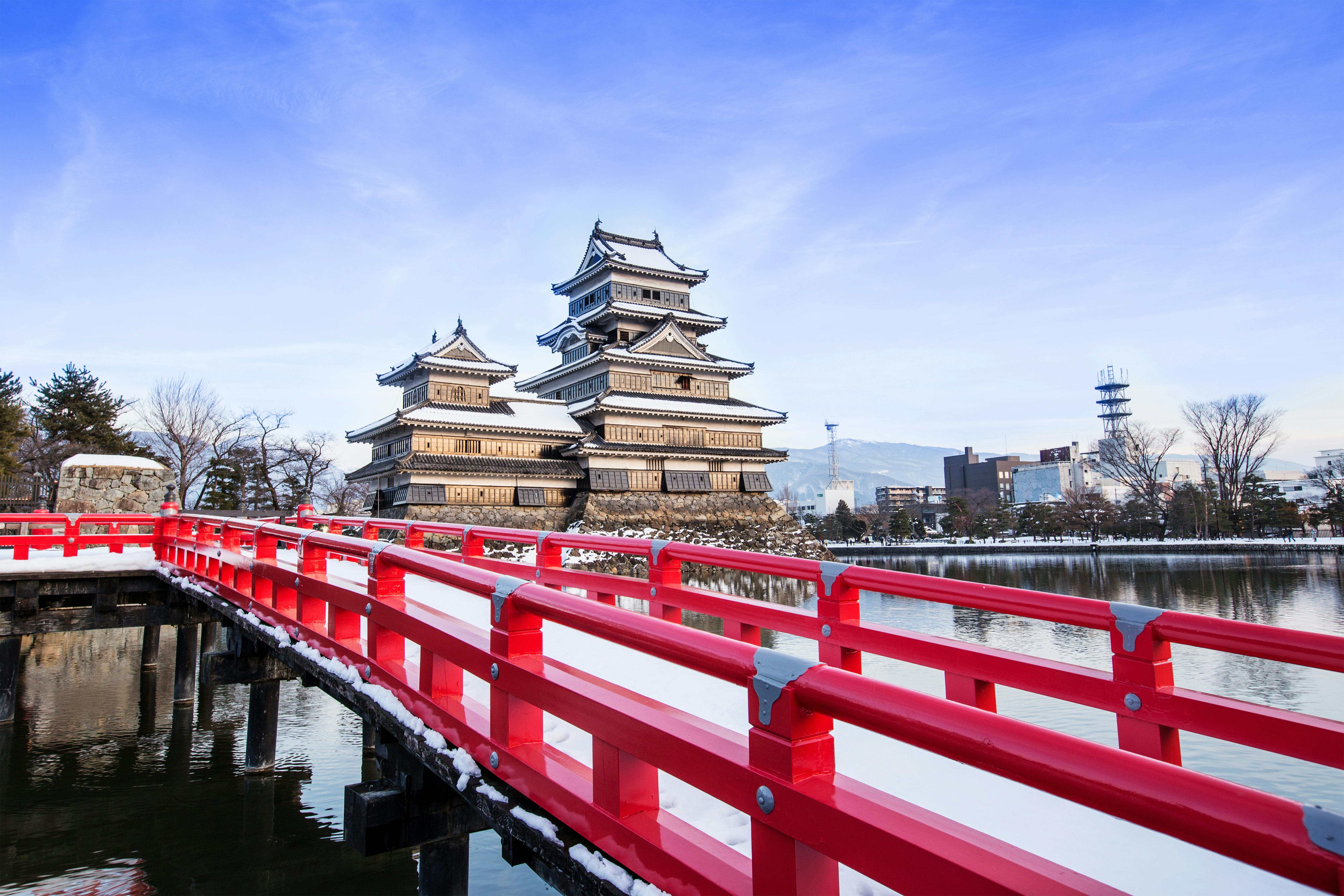 A bright red bridge leads to Matsumoto Castle, a historic Japanese castle with black and white towers, surrounded by a moat under a clear blue sky in winter.