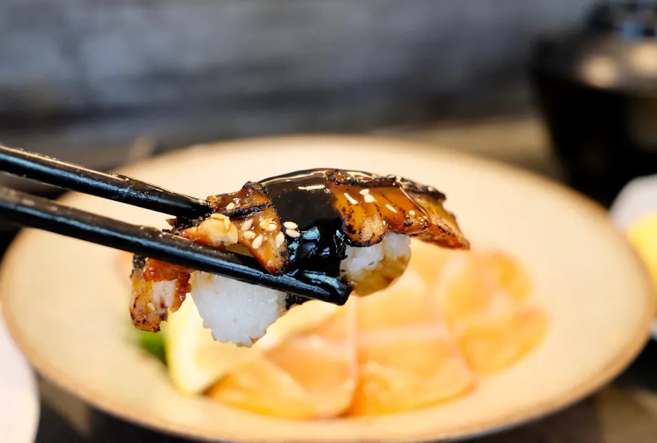 特写镜头展示一双筷子夹着一块覆盖有鳗鱼（烤鳗鱼）并淋上深色酱汁，可能是照烧酱的寿司，背景模糊处是盘子上更多的寿司。